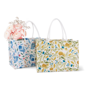 ALT Two large printed canvas tote bags with white webbing handles, one featuring a blue botanical floral print and the other a yellow floral print, with pink roses placed inside.