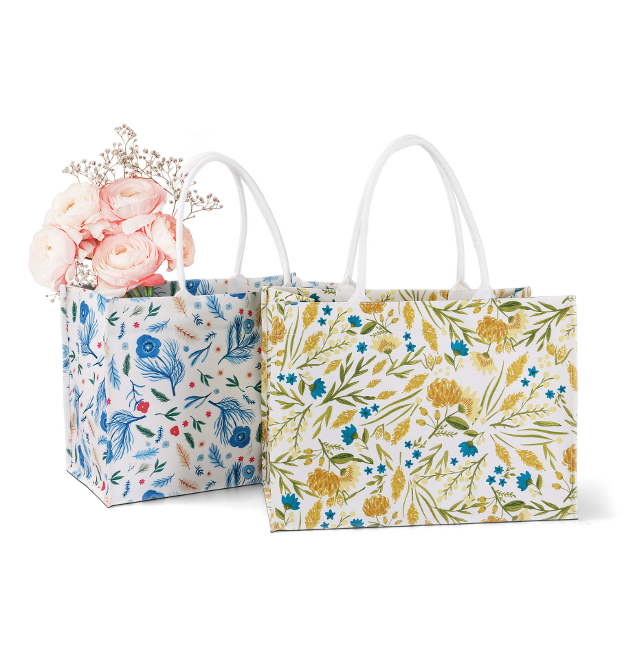ALT Two large printed canvas tote bags with white webbing handles, one featuring a blue botanical floral print and the other a yellow floral print, with pink roses placed inside.