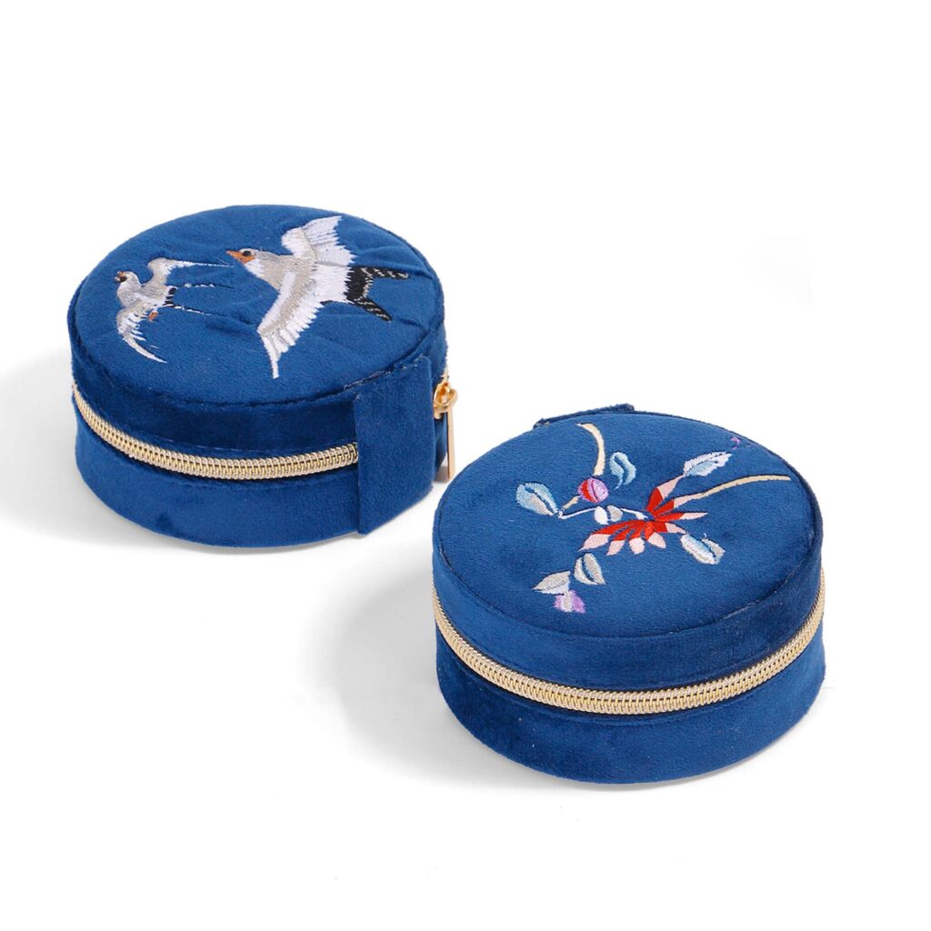 ALT Two round blue velvet jewelry boxes with gold zipper trim, one featuring embroidered white birds and the other with a floral branch design in red, white, and purple.