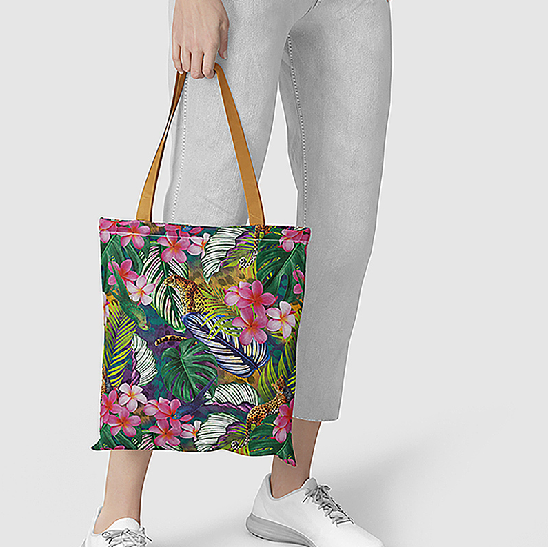 ALT A person holding a tropical jungle print canvas tote bag with brown leather handles, featuring pink plumeria flowers, green palm leaves, and leopards.