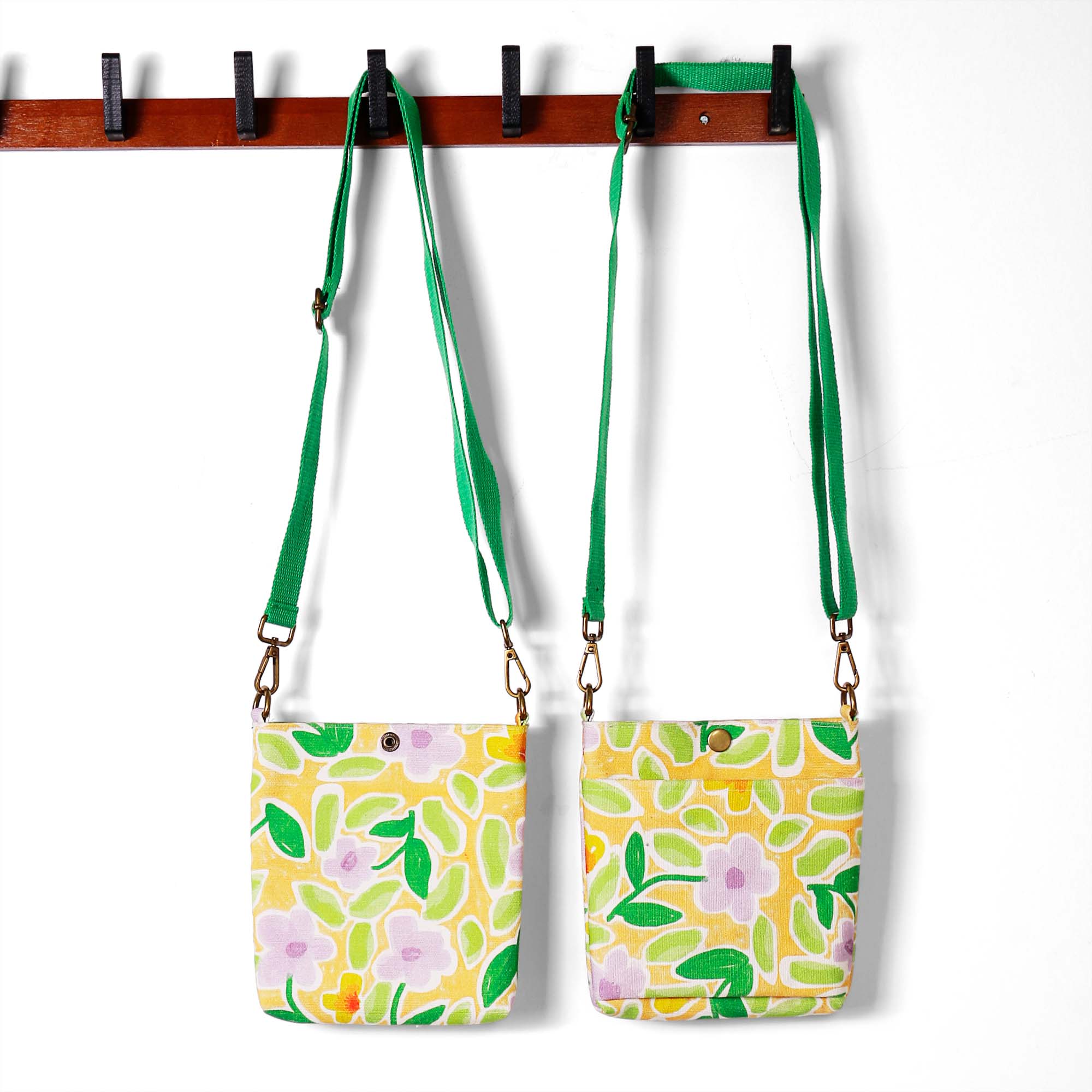 ALT Two floral canvas crossbody bags hanging on a wooden rack, one with a green webbing strap and the other with a lavender strap, featuring a yellow background with green leaves